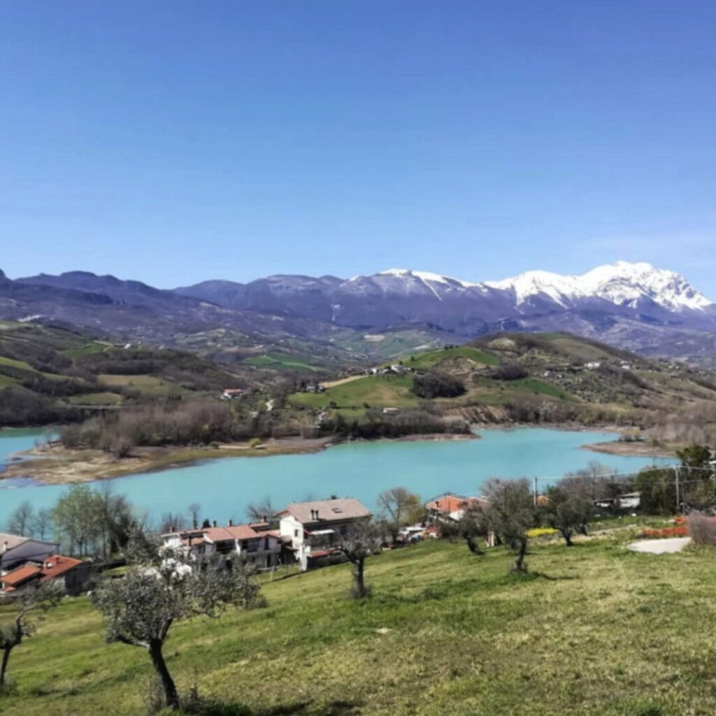 Turquoise lake nestled in green hills of Abruzzo, Italy, with olive trees in the foreground, small houses, and snow-capped mountains under a clear blue sky – a serene hidden gem for nature lovers.
