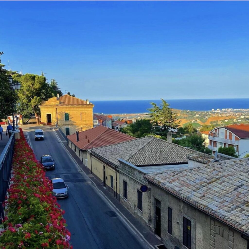 Elevated view of a flower-lined street in an Abruzzo coastal town, with red-tiled roofs, passing cars, and distant Adriatic Sea horizon under sunny skies – perfect for relaxed escapes in Italy's undiscovered regions