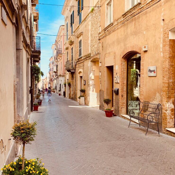 Calle peatonal soleada en el centro histórico de Guardiagrele, flanqueada por edificios ocres, plantas en macetas y un banco de hierro forjado