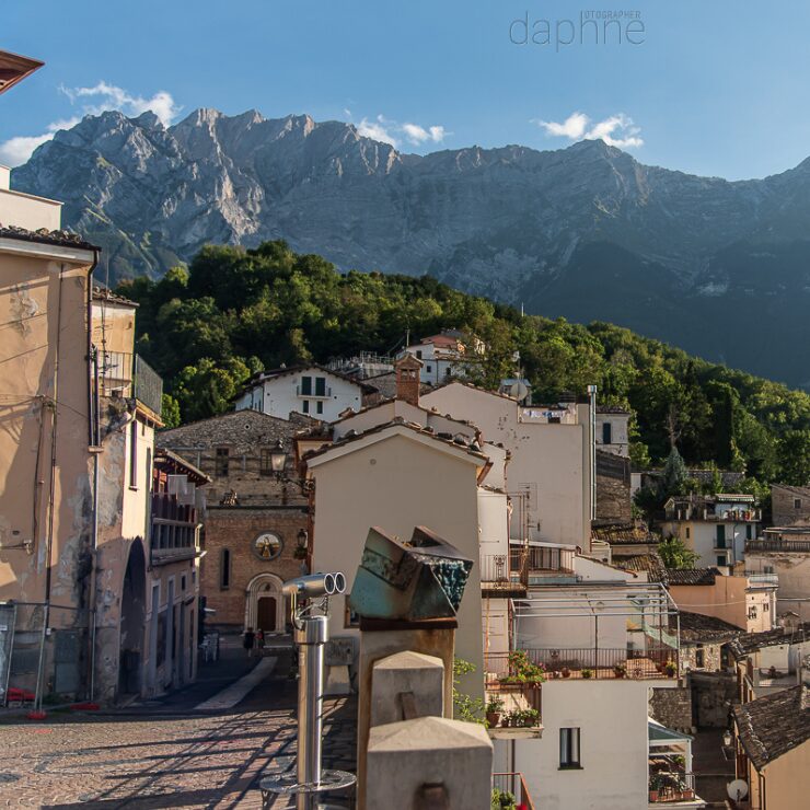 Vista panorámica del centro histórico de un pueblo abrucés con tejados de piedra, una plaza con binoculares panorámicos y las escarpadas cumbres rocosas de la Maiella al fondo bajo un cielo azul. Vista panorámica del centro histórico de un pueblo abrucés con tejados de piedra, una plaza con binoculares panorámicos y las escarpadas cumbres rocosas de la Maiella al fondo bajo un cielo azul.
