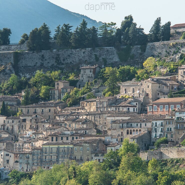 Vista a distancia de un pueblo medieval abrucés en la ladera de una colina, con casas de piedra apiladas y murallas históricas visibles en la parte superior, rodeadas de vegetación.