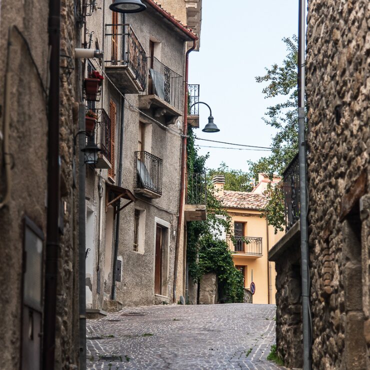Callejuela empedrada de un pueblo abrucés encuadrada entre muros de piedra, con balcones de hierro forjado, una farola clásica y una fachada amarilla al fondo entre vegetación.