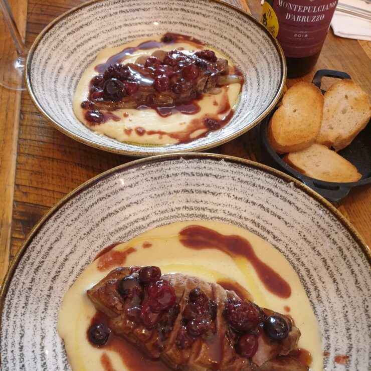 Due piatti di secondo con carne in salsa di frutti di bosco su crema, accompagnati da pane tostato e una bottiglia di Montepulciano d'Abruzzo