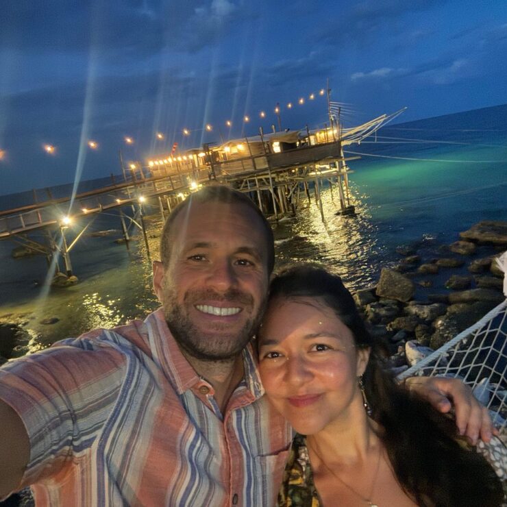 Una pareja sonriendo con un trabocco iluminado sobre el mar Adriático de fondo por la noche en Abruzzo