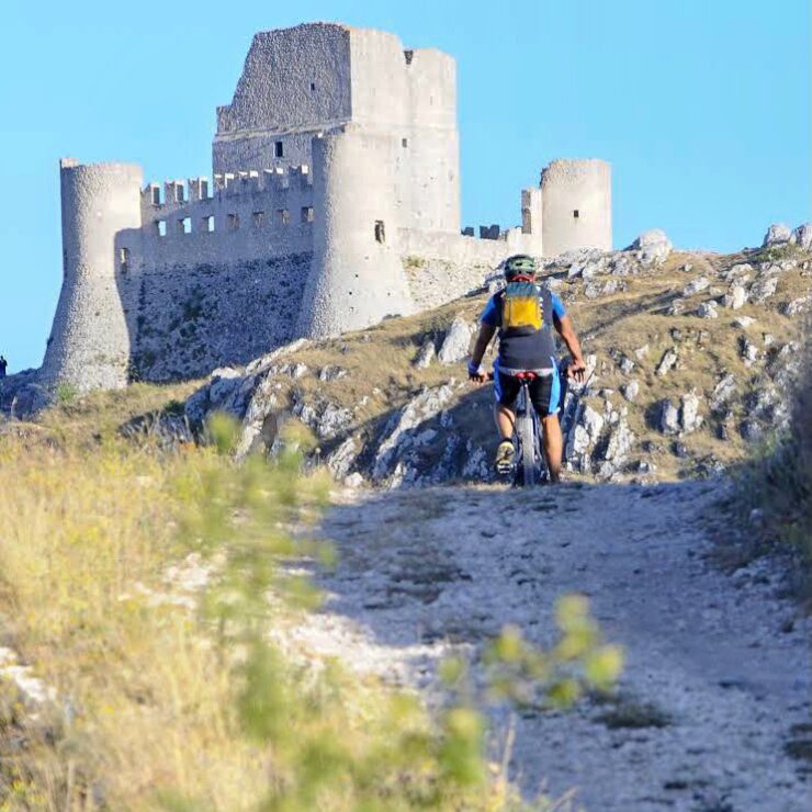 Un ciclista di montagna che percorre un sentiero sterrato verso le rovine del castello medievale di Rocca Calascio sugli altopiani abruzzesi