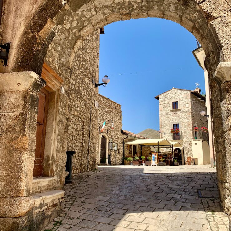 Cobblestone Medici Square in the medieval village of Santo Stefano di Sessanio, Abruzzo, Italy, surrounded by stone archways and historic buildings under a clear sky, with the village's entrance portal bearing the Medici coat of arms.