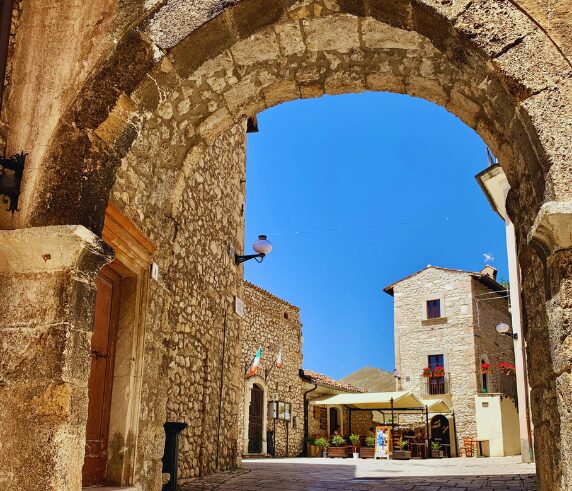 Cobblestone Medici Square in the medieval village of Santo Stefano di Sessanio, Abruzzo, Italy, surrounded by stone archways and historic buildings under a clear sky, with the village's entrance portal bearing the Medici coat of arms.