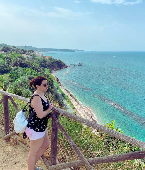 Idania leaning on a wooden fence, gazing at the turquoise Adriatic Sea and a trabocco fishing platform along the lush Costa dei Trabocchi in Abruzzo, Italy.