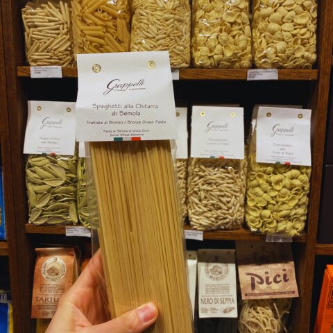 A hand holding a packet of spaghetti alla chitarra in front of a wooden shelf stocked with various shapes of artisan Italian pasta