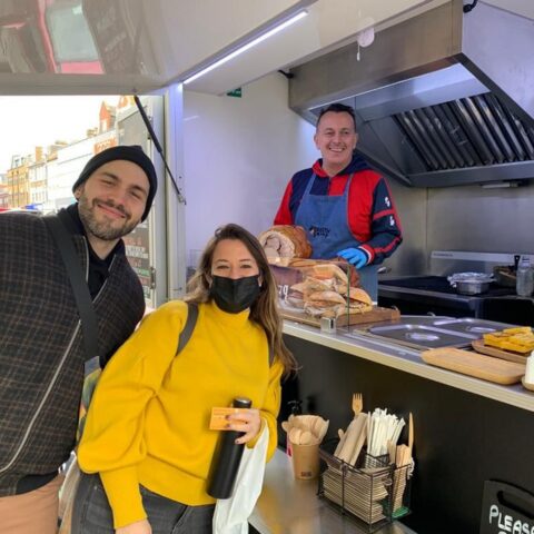 Two people smiling at an outdoor food stall being served by a chef, at a street market in London