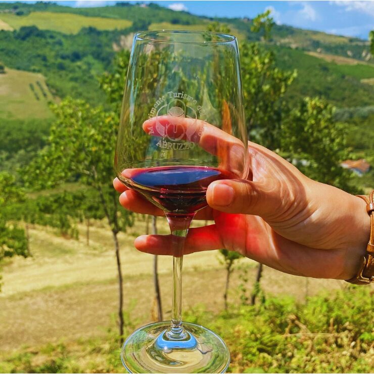 Una mano sosteniendo una copa de vino grabada con el logo de turismo de Abruzzo, con viñedos y colinas verdes al fondo