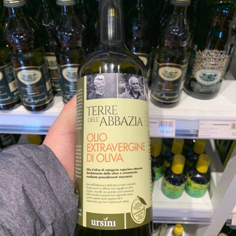 A hand holding a bottle of Terre dell'Abbazia extra virgin olive oil by Ursini in a supermarket aisle